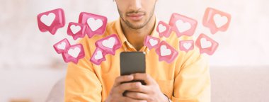 Bir adam kanepede oturuyor, elinde bir akıllı telefon tutuyor, ekrana bakıyor. Sosyal medya tebligatlarını hatırlatan bir dizi pembe kalp simgesi etrafında uçuşuyor..