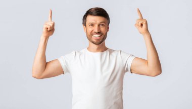 Beyaz tişörtlü, gülümseyen bir adam her iki işaret parmağını da yukarı kaldırıp doğrudan kameraya bakar..