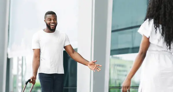 stock image Black man with luggage is standing in a modern buildings glass-walled entrance, smiling and greeting a woman who is walking towards him.