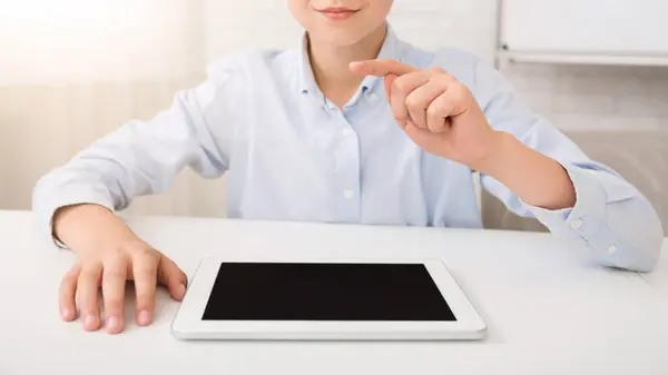 stock image A young boy sits at a white desk in a bright room. He is wearing a light blue button-down shirt and is using a digital tablet with a black screen. He is pointing with his index finger at the tablet.