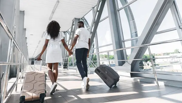 stock image Back view of black couple walks hand-in-hand through an airport terminal, each pulling a suitcase behind them. They are dressed casually and seem to be happy and excited to be traveling, copy space