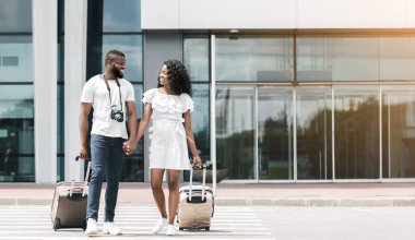 Mutlu siyah bir çift havaalanı terminalinin önünde el ele yürür, her biri arkalarında yuvarlanan bir bavul taşır. İkisi de gülümsüyor ve birbirlerine bakıyorlar, birlikte vakit geçiriyorlar..