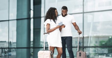 Mutlu bir siyah çift havaalanının dışında uçuş kartlarını kontrol ediyor. İkisinin de elinde bavul var. Gülümsüyorlar ve birbirlerine bakıyorlar. Tatillerine başlamak için heyecanlı görünüyorlar..