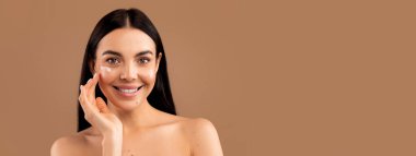 Neşeli, çıplak, uzun saçlı, esmer, yüzünde tebessüm olan, genç, parlak cilt için yüz kremi kullanan, bej renkli stüdyo arka planında izole edilmiş, kopyalama alanı olan bir kadın.