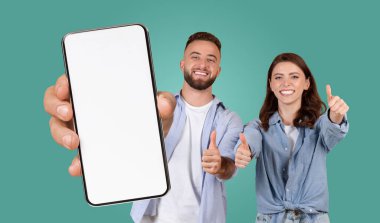 Genç bir adam ve kadın elinde boş ekranlı akıllı bir telefon tutuyorlar ve kameraya bakarken başparmaklarını kaldırıyorlar. Parlak yeşil bir arkaplanda duruyorlar.