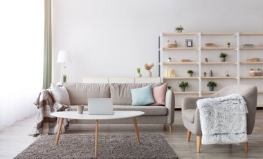 Evden iş, serbest çalışma ve Covid-19 salgını sırasında uzaktan çalışma. Yastıklı gri kanepe, dizüstü bilgisayarlı masa ve bir fincan sıcak içecek, raflar ve modern oturma odasında battaniyeli koltuk.