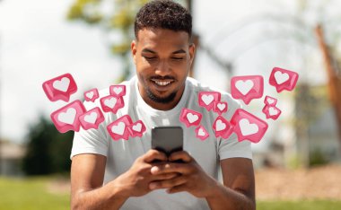 Genç siyah bir adam parkta oturuyor, akıllı telefonuna bakarken gülümsüyor. İki eliyle telefonu tutuyor ve etrafında birden fazla kalp şeklindeki ikonlar dolaşıyor.