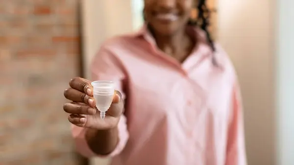 stock image African American woman, wearing a pink shirt, holds a menstrual cup in her hand. She is smiling and looking toward the camera, cropped