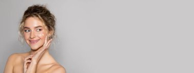 Neşeli, çıplak, uzun saçlı, yüzünde gülümseme olan, genç, pürüzsüz bir cilt için yüz kremi kullanan, gri stüdyo arka planında izole edilmiş, uzun saçlı, sarışın, milenyum kadını.