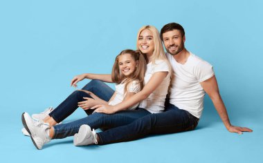 Üç kişilik bir aile açık mavi bir arka planda oturuyor, gülümsüyor ve kameraya bakıyor. Hepsi gündelik kıyafetler giyiyor.