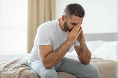 Beyaz tişört ve gri pantolon giyen bir adam bir yatağın kenarında oturuyor. Yüzünde elleri var ve aşağıya bakıyor, derin düşüncelere dalmış gibi görünüyor..