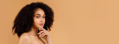 Genç, güzel, siyah bir bayan pürüzsüz tenine dokunuyor. Başarılı yüz derisi rutininin tadını çıkarıyor. Havluyla poz veriyor, bej arka planda kameraya bakıyor.