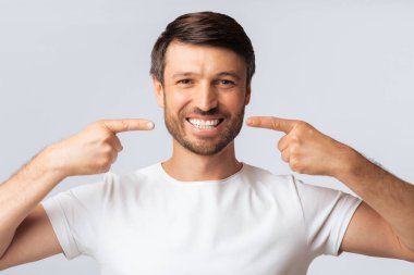 Sakallı bir adam her iki işaret parmağıyla dişlerini gösterir. Beyaz bir tişört giyiyor ve beyaz bir arkaplanda duruyor..