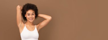Gülümseyen bin yıllık Afro-Amerikan kıvırcık saçlı, beyaz tenli kadın kameraya bakıyor, boş zamanın tadını çıkarıyor, kahverengi arka planda, stüdyoda, kopyalama alanında izole edilmiş.
