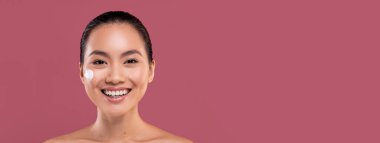 Güzel yüzünde nemlendirici kremi olan neşeli, çekici, üstsüz, genç Asyalı kadın yeni güzellik ürünlerini test ediyor, kameraya gülümsüyor, mor stüdyo arka planı, kopyalama alanı. Yüz bakımı kavramı