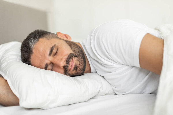 A man with a beard is lying on his side in a bed, sleeping soundly. His eyes are closed, and his face has a relaxed expression. He is wearing a white t-shirt and is covered with a white blanket.
