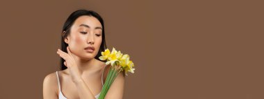 Kahverengi arka planda sarı nergis çiçekleriyle poz veren genç, güzel Japon bayan, stüdyo çekimi. Güzellik ve cilt bakımı, yaşlanma karşıtı tedavi, spa prosedürü, reklam ve teklif, kopyalama alanı