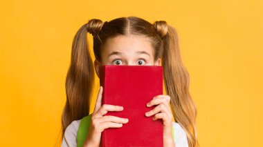 Okula dönüş kaygısı. Korkak ilkokul öğrencisi kız sarı stüdyo arka planında kitabın arkasına saklanıyor. Boş Alan
