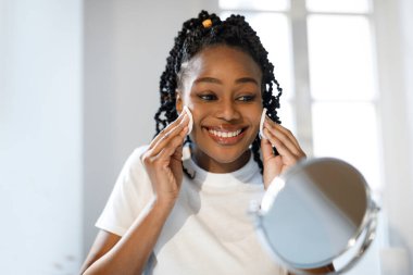 African American young woman with braids smiles as she cleanses her face with cotton pads, looking at her reflection in a round mirror. clipart