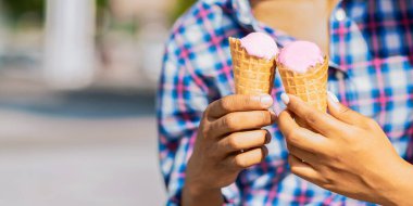 İçinde pembe dondurma olan iki dondurma külahı tutan birinin yakın plan görüntüsü. Kişi ekose bir gömlek giyiyor ve güneşli bir günde dışarıda. Arkaplan bulanık ve bulanık.