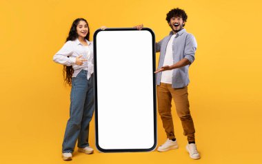 Cool mobile offer. Happy indian couple pointing at big cellphone with white screen for mockup, demonstrating free space for phone application or website design, yellow background