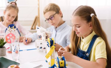 Üç genç öğrenci bir sınıf ortamında bir masada oturuyorlar, robotlar üzerinde çalışıyorlar. İşlerine yoğunlaşıyorlar ve etkinlikten zevk alıyor gibi görünüyorlar..