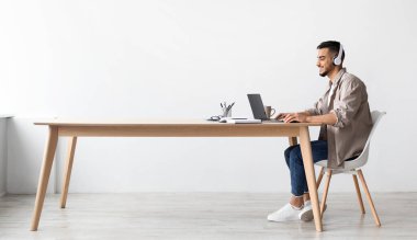 Neşeli Ortadoğulu serbest çalışan, PC 'de çalışan, kablosuz kulaklıklarla müzik dinleyen, yeni proje için araştırma yapan, masa başında oturup uzaktan izlemenin keyfini çıkaran,