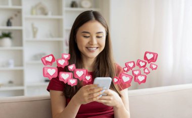Çinli genç bir kadın kanepede oturuyor ve akıllı telefonunu kullanırken gülümsüyor. Çevresini çevreleyen birçok dijital pembe kalp simgesi var. Sevdikleri ya da sosyal medya bağlantılarını temsil ediyorlar..