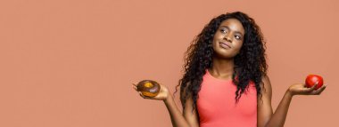 Uzun kıvırcık saçlı, pembe kolsuz tişörtlü, şeftali renkli bir arka planın önünde duran Afro-Amerikalı genç bir kadın. Bir elinde jöleli bir çörek, diğer elinde kırmızı bir elma tutuyor.