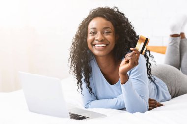Neşeli Afrikalı kadın evde yatakta yatıyordu, online alışveriş, dizüstü bilgisayar kullanarak ve kredi kartı tutarak, kopya alanı