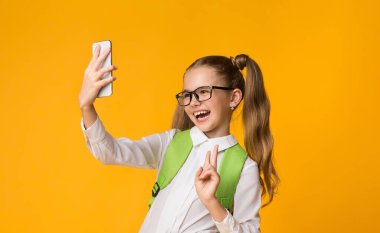 Okul Selfie'si. Mutlu Schoolgirl Smartphone Taking Self-Portrait Gesturing Zafer İşareti Kullanarak Sarı Studio Arka Plan. Kopyalama Alanı