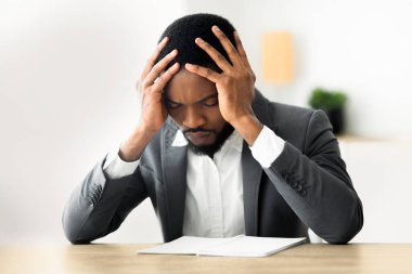 Takım elbiseli bir işadamı, kafası ellerinin arasında bir masada oturur ve evrak işlerine bakar..