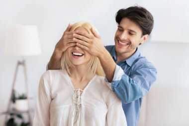 Bir erkek elleriyle gözlerini kapatarak kız arkadaşına sürpriz yapar. Beklenti içinde gülümsüyor.