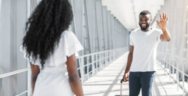 Bavullu Afrikalı Amerikalı bir adam havaalanında bir kadını selamlıyor. Adam gülümsüyor ve elini sallarken kadın ona bakmıyor..