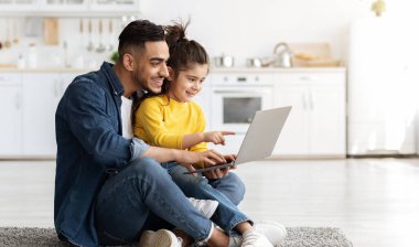 Küçük kızı ile mutlu bir Arap adam evde dizüstü bilgisayar kullanıyor, Orta Doğulu baba ve kız çocuğu online alışveriş için bilgisayar kullanıyor ya da mutfakta yerde dinlenirken interneti tarıyor.