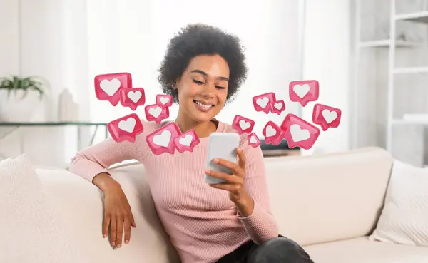 stock image A young Black woman with curly hair is sitting on a white couch in a living room, smiling as she looks at her smartphone. She is wearing a light pink sweater and has her arm resting on the couch