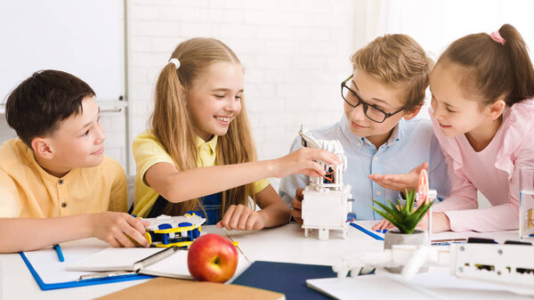 Четверо детей, двое мальчиков и две девочки, работают вместе над проектом робототехники в классе. Они все улыбаются и, кажется, занимаются поставленной задачей.