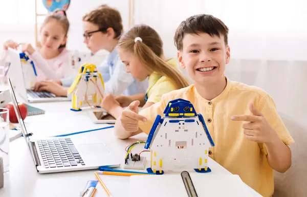 stock image A young boy sits in a classroom, beaming with pride as he points to a robot he built. He is surrounded by other students who are also engaged in STEM activities.