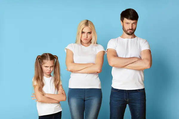 stock image A family of three stands in front of a light blue background with their arms crossed. The young girl has a frustrated expression on her face. In the middle is the mother appears to be annoyed.