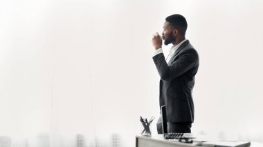 Takım elbiseli bir adam pencerenin yanında durur ve dışarı bakarken kahve içer..