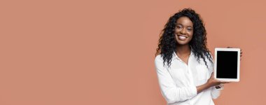 Uzun kıvırcık saçlı genç siyah bir kadın şeftali renkli bir arka planın önünde beyaz bir tablet tutarken gülümsüyor. Tablet ekranı siyah ve kadın beyaz düğmeli gömlek giyiyor..