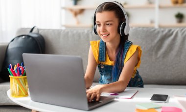 Portrait of little girl in wireless headset using laptop, studying online at home, interested happy student typing on keyboard looking at pc screen, watching webinar, online course, doing homework