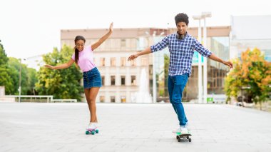 Genç bir Afro-Amerikalı erkek ve kız şehir parkında kaykay yapıyor, gülümsüyor ve eğleniyorlar. İkisi de gündelik kıyafetler giyiyorlar ve adamın kolu açık..