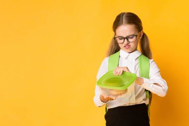 Disgusted Schoolgirl Looking At Sandwich In Lunchbox Over Yellow Studio Background. School Lunch And Gluten Intolerance. Copy Space clipart