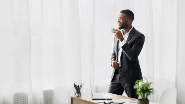 Takım elbiseli gülümseyen bir işadamı, elinde kahve fincanı, pencerenin yanında durur ve pencereden dışarı bakar..