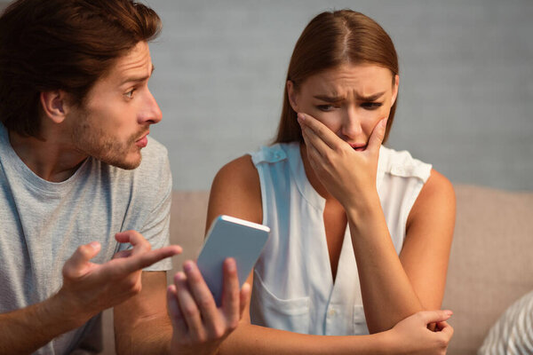 Infidelity. Jealous Husband Showing His Cheating Wife Her Phone Demanding Explanation Sitting On Sofa Indoor. Selective Focus