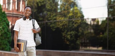 Sırt çantası ve çalışma kitapları olan mutlu gülümseyen Afrikalı Amerikalı öğrenci, kameraya bakıyor ve gülümsüyor.