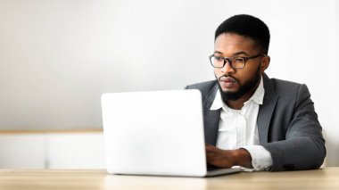Ofiste dizüstü bilgisayar üzerinde çalışan kendine güvenen Afro-Amerikan işadamı, ciddi yüz ifadesi ile ekrana bakıyor, fotokopi alanı ile panorama