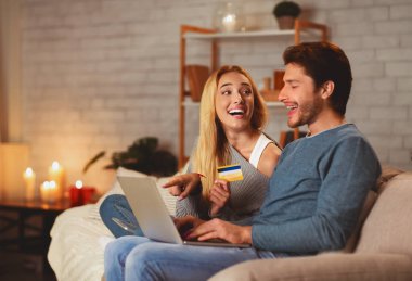 E-Ticaret ve İnternet Alışverişi. Heyecanlı çift dizüstü bilgisayar ve kredi kartı tutuyor. Kapalı koltukta oturmuş internetten bir şeyler alıyorlar.