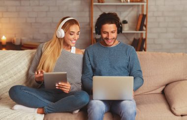Modern Yaşam Tarzı. Gülümseyen Çift Akşam Evde Kanepede Oturan Kulaklık Giyen Dizüstü Ve Tablet Bilgisayarları Kullanarak